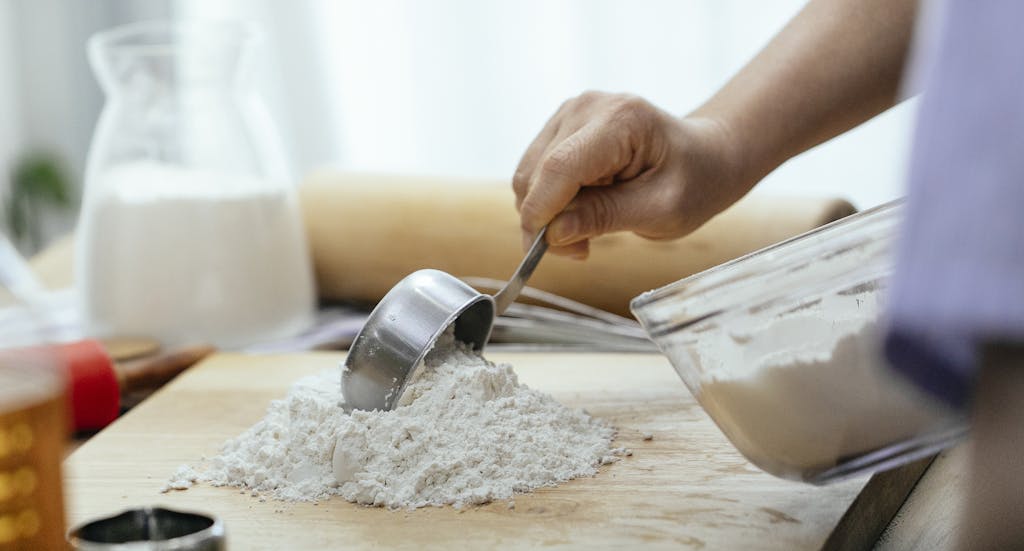 Crop adult woman adding flour in a batter bowl on wooden cutting board