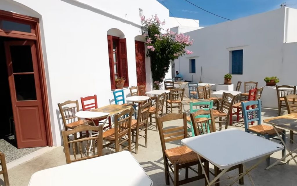 multiple colors chairs on a white stone terrace in front of a white restaurant with brown wooden shudders and door, in Greece