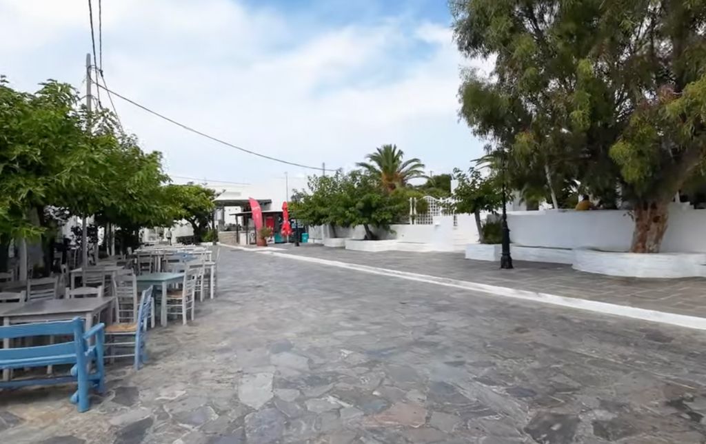 large pedestrian stone paved road on a greek island with blue chairs and green trees alongside the road