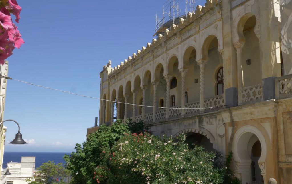 ancient villa with archways and sculptures, green bushes in front of it, sea in the background