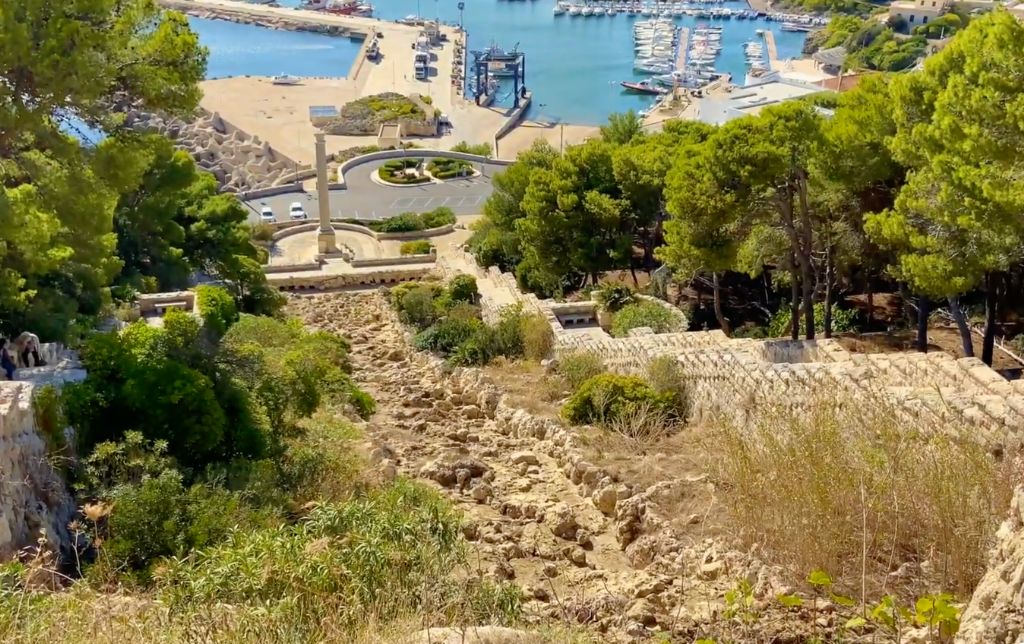 ancient ruins going downhill with trees on both sides; gulf with boats in the background