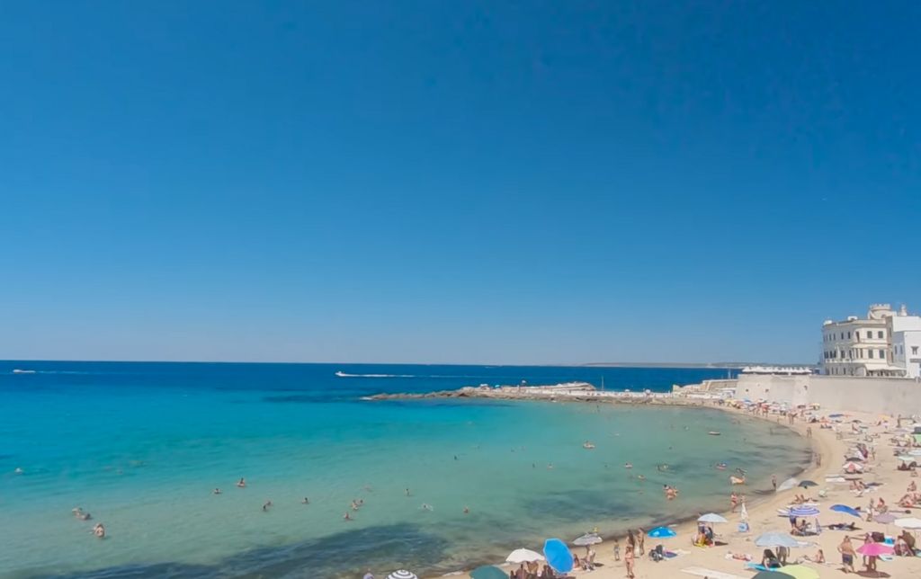 beach with white sand and a big building in the background; colourful umbrellas and many people on the beach and in the water