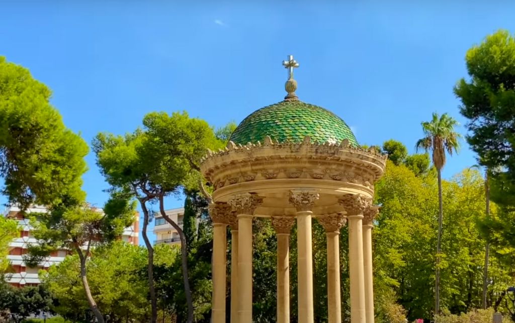 Christian monument with green round roof, surrounded by trees and buildings in the background