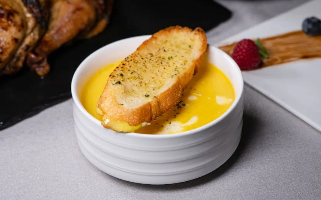 a garlic bread resting on a white soup bowl on a white table. in the background a blueberry and a strawberry can be seen