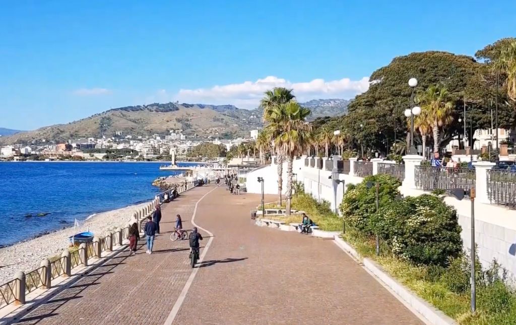 waling path along the beach, people riding bikes or walking on the path, big old trees planted on the right side of the walking path