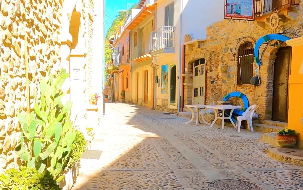 stone road, stone building on the right and left on the road and two white tables with one white chair