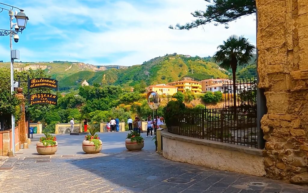 wide stone road exiting the town towards the green hills, people standing in a vista point, watching the hills