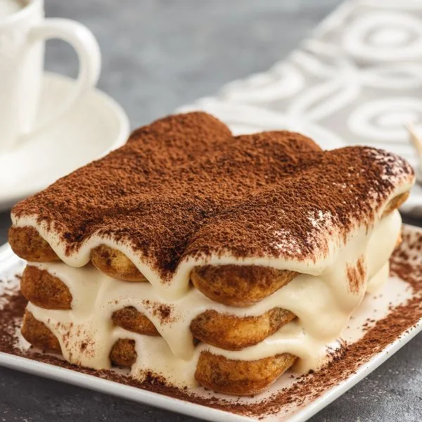 tiramisu on a white square plate on a blue grey table near a cup of coffee