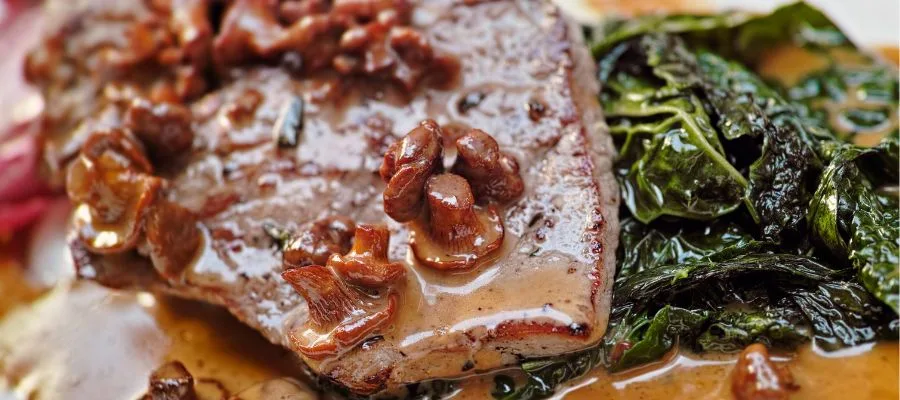 detail of a a piece of veal marsala with green cooked spinach and mushrooms, sitting on a beige plate