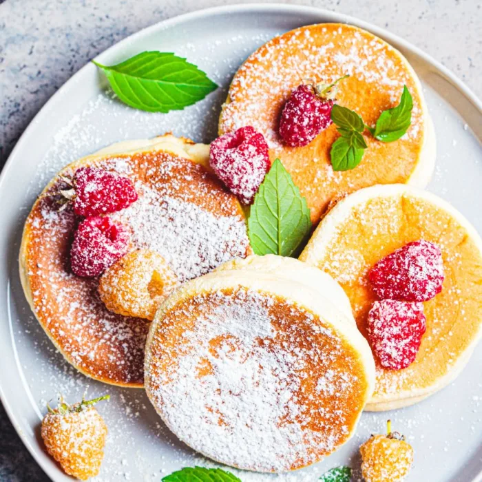 four japanese pancakes on a white plate, with sugar and raspberries on top