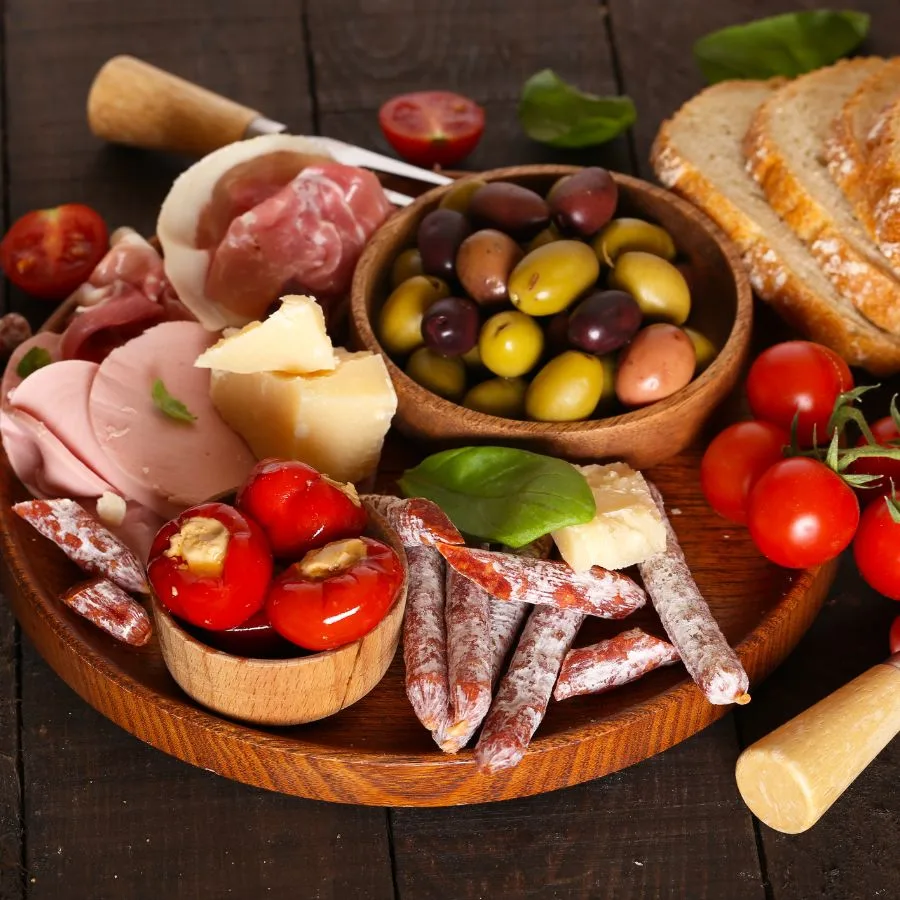 olives in a bowl, tomatoes filled with cheese in another bowl, tomatoes fresh, cheese, salami and ham, all on a wooden board