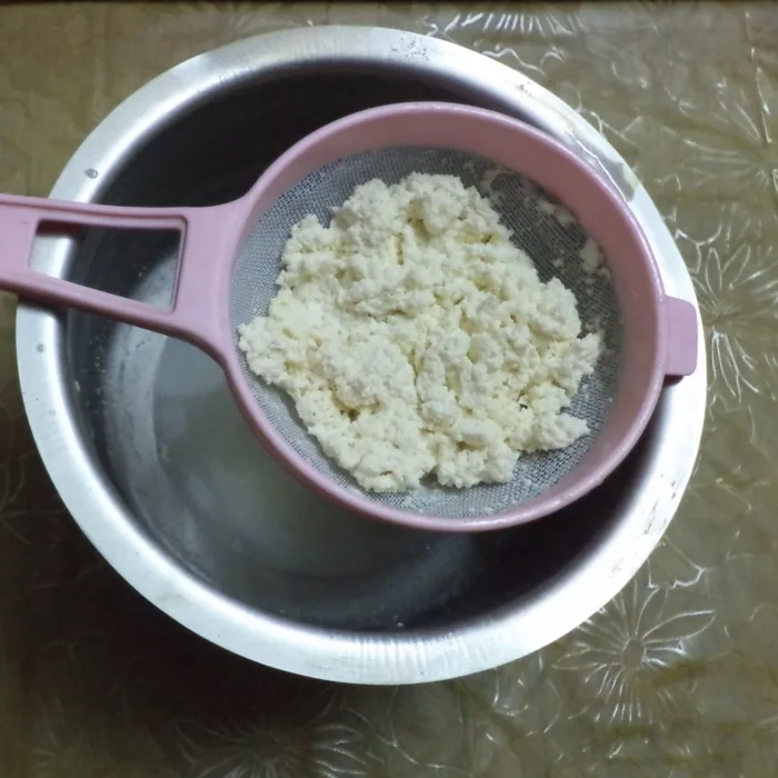 a pink sieve sitting on a stainless steel bowl, holding cheese, on a wooden table