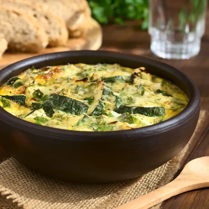 Zucchini-Mint Frittata in a brown ceramic bowl on a wooden table near bread slices and a glass of water and parsley and a wooden spoon