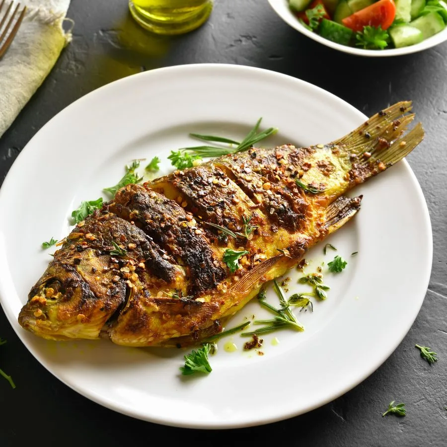 grilled fish with lemon and herbs sitting on a white plate on a grey stone table
