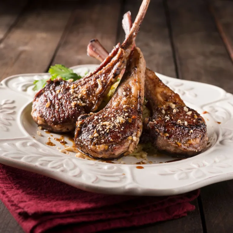 3 grilled lamb chops on a white plate sitting on a wooden table near a red kitchen towel