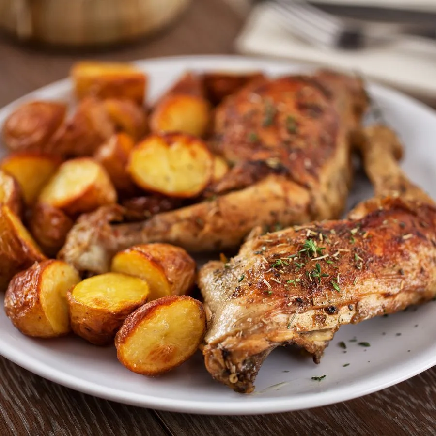 2 legs of crispy chicken with raosted potatoes on a white table on a wooden table near a fork placed on a white kitchen towel