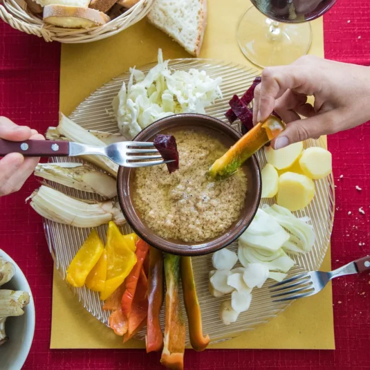 bagna cauda in the middle in a brown pot surrounded by bell peppers, cream cheese, people dipping in it on a red table